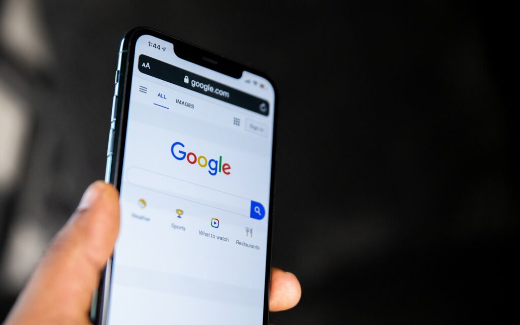 Hand hält Smartphone mit Google-Startseite, über die sich der Google AI Mode aufrufen lässt